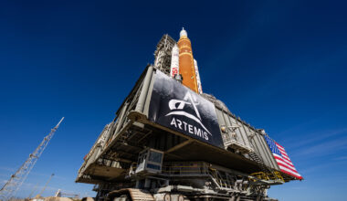 NASA's Artemis II SLS rocket - with its distinctive butterscotch orange-brown color- and Orion spacecraft are on top of a massive platform. The platform has a banner with the Artemis logo on it on the left and an American flag on the right. The rocket and platform are moved by an immense machine called the crawler-transporter, which has treads like a tank. The view is from the ground up, so the deep blue sky is the background for most of this picture.