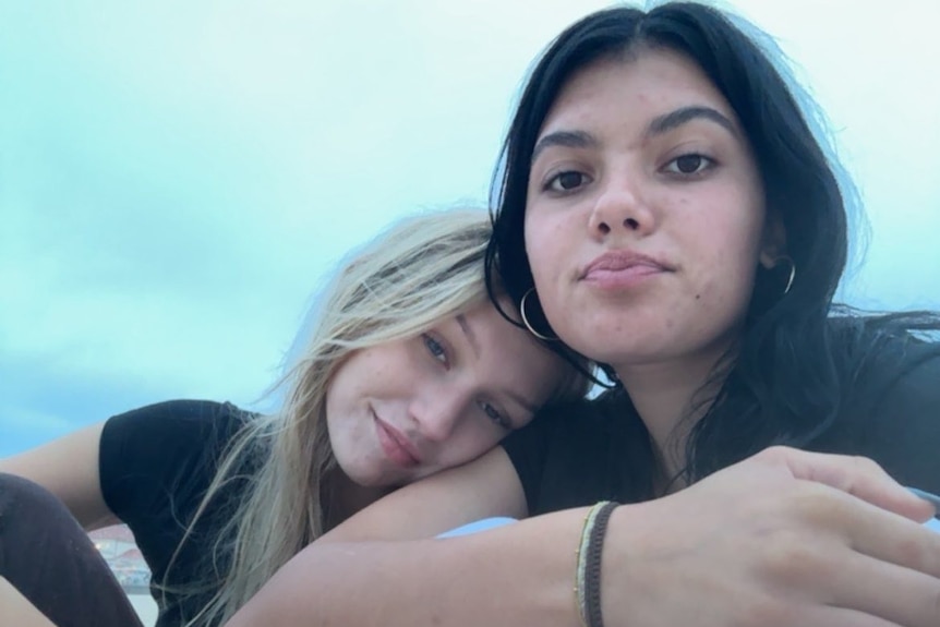 A blonde girl resting her head on the shoulder of a  dark-haired girl.