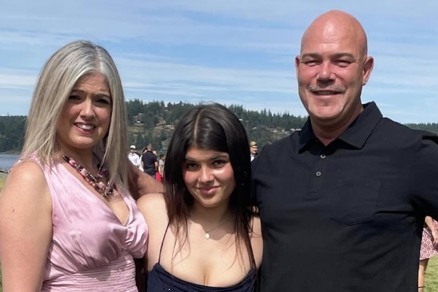 A yuoung, dark-haired woman arm in arm with a blonde woman and a smiling man in an outdoor area.