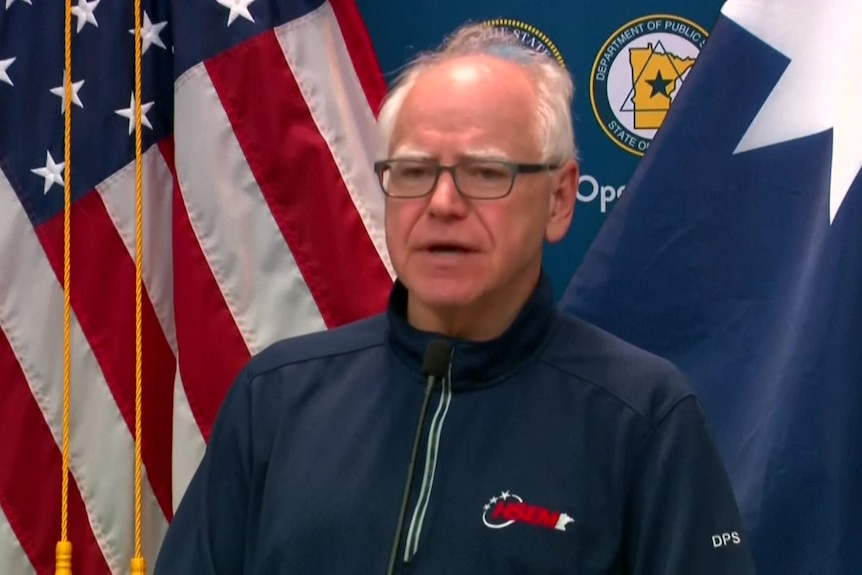 A man with white hair stands in front of a US flag during a press conference. 