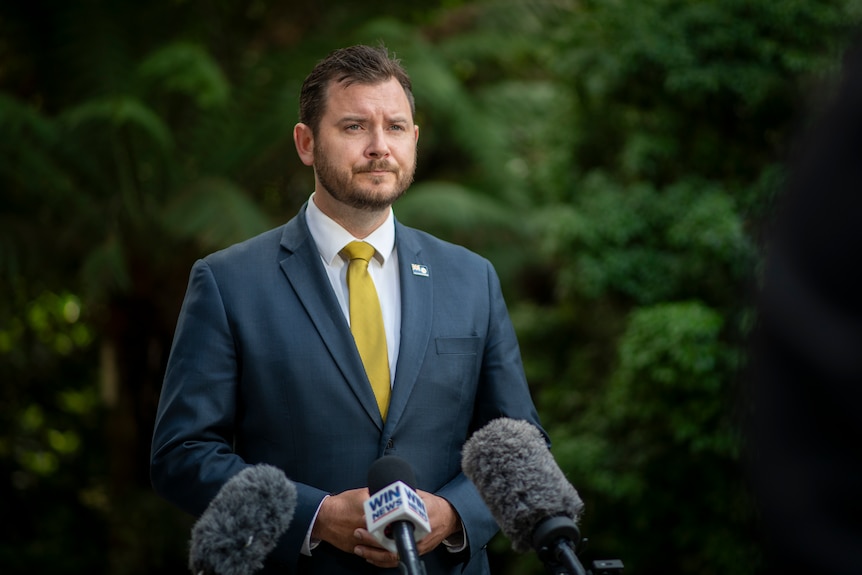 A man with dark facial hair, a navy suit and yellow tie stands over microphones with soft green rainforest in the background.