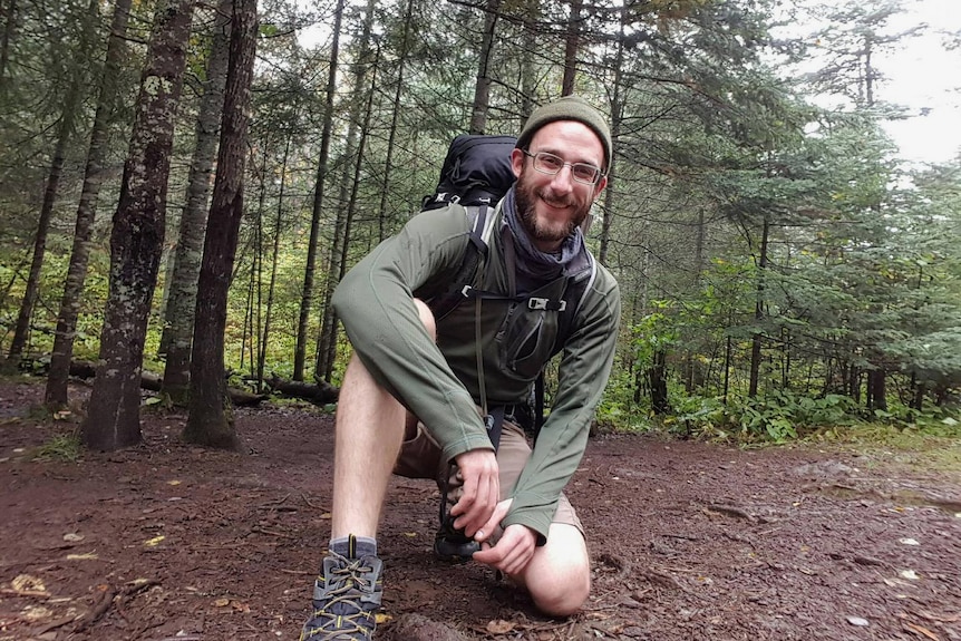 A man with backpack and hiking boots, poses by some trees with one knee on the ground.