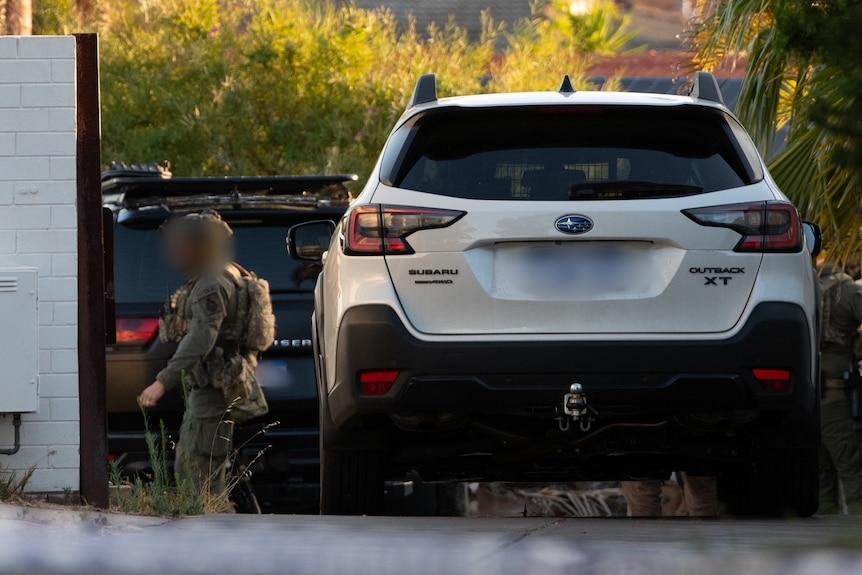 A police officer in camouflage gear stands outside a home with cars alongside.