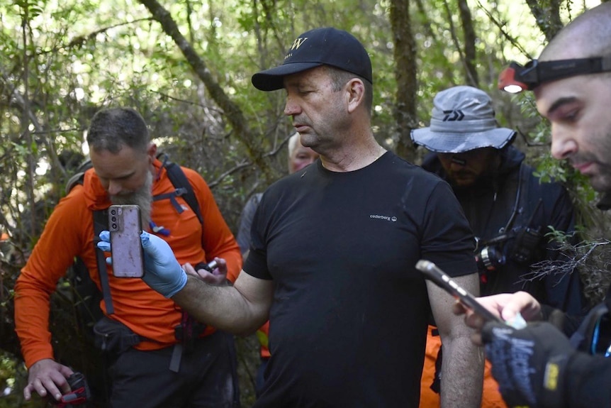 Ken Gamble wears a black t-shirt and black cap, and holds a mobile phone in a gloved hand as three other people look on.