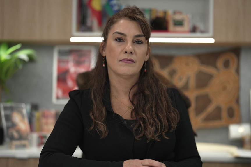 A woman with brown wavy hair is wearing a black blazer and sitting in an office