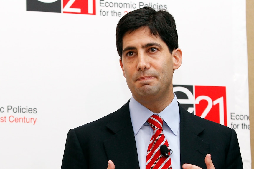 A photo of a man in his 40s with short black hair, wearing a red tie and black suit.