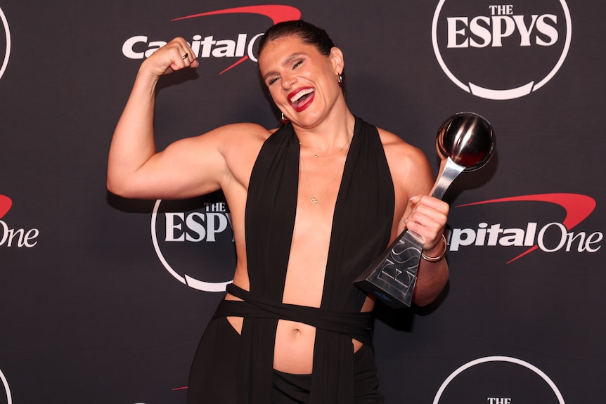 USA women's rugby player Ilona Maher is wearing a black cutout dress and flexes her muscles, smiling, while holding a trophy