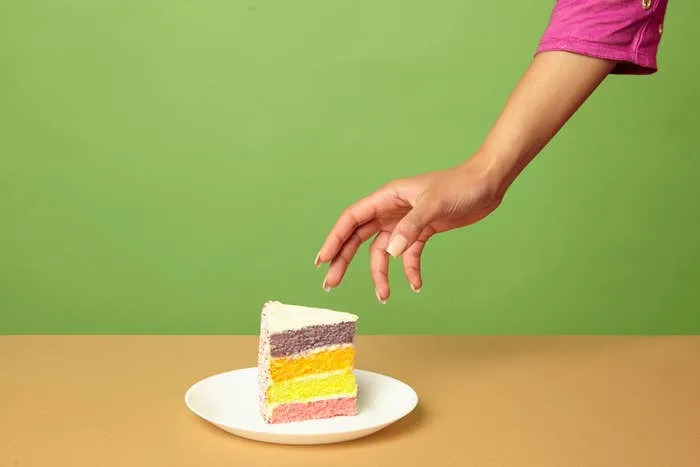 A hand reaches for a slice of rainbow layered cake on a white plate against a plain background