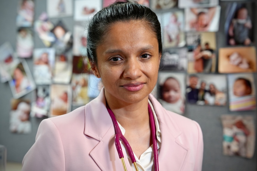 A woman with her hair up in a pink suit looks directly at the camera. 