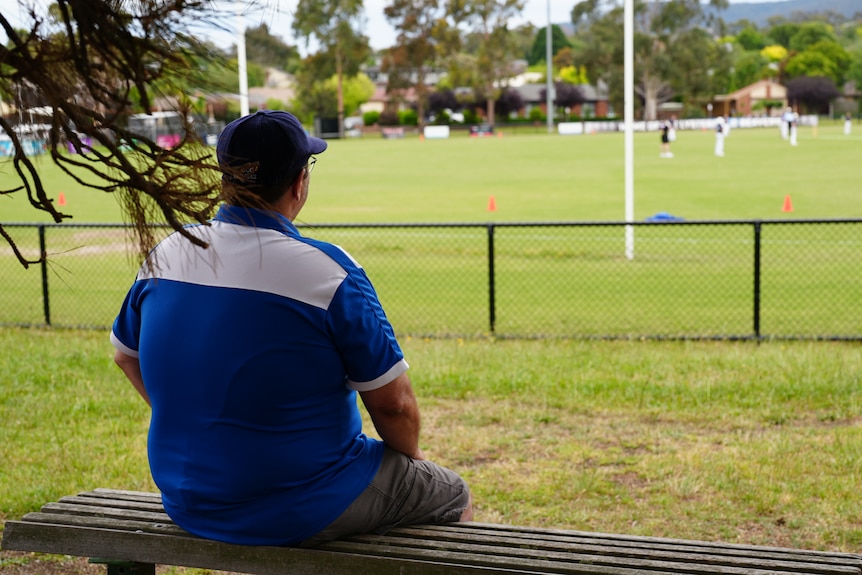 A man sits on a bench watching a game of cricket.