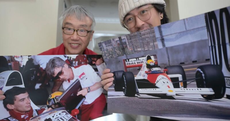 Two men smile at the camera while holding large photos of Formula 1 scenes, including a driver signing an autograph and a racing car on the track.