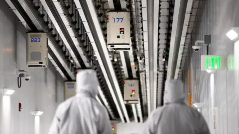 Bloomberg via Getty Images Employees wearing cleanroom suits walk beneath Automated Material Handling Systems (AMHS) vehicle robots moving along tracks on the ceiling inside the GlobalFoundries semiconductor manufacturing facility in Malta, New York, U.S., on Tuesday, March 16, 2021. 
