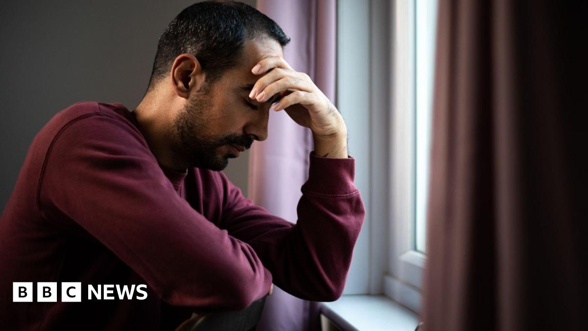 Man in maroon jumper with his head in his hands, sat by his window. He has dark hair and dark facial hair.