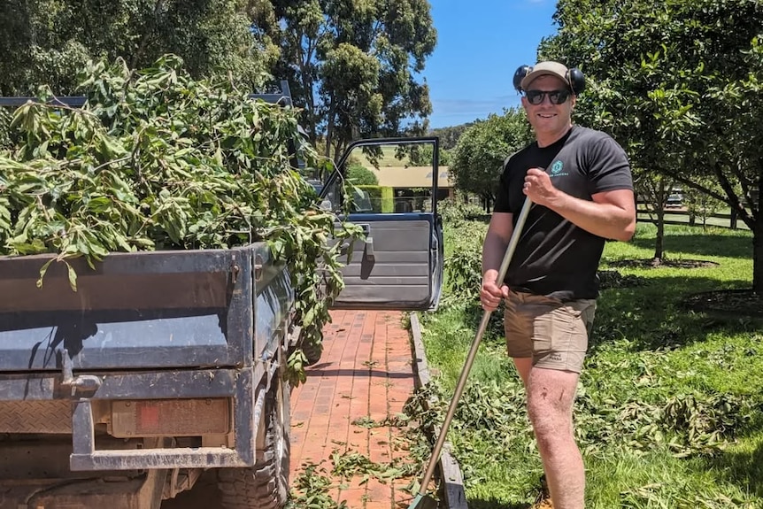 A man holding a rake. He is standing beside a ute filled with dried leaves and branches.