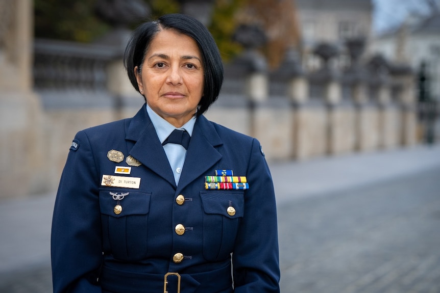 A woman in a military uniform standing near a large fence looks at the camera with a neutral expression