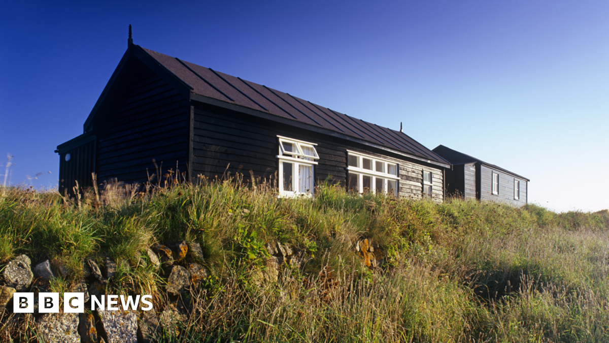 A wide shot of the radio station. It is a brown single-storey hut surrounded by grass.
