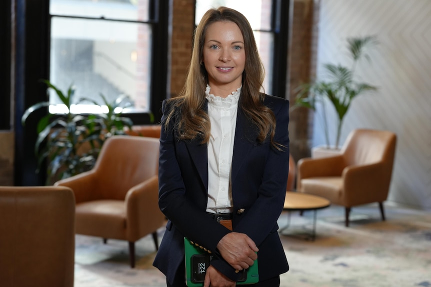 A woman in a suit in a room with brown chairs and a few indoor plants.