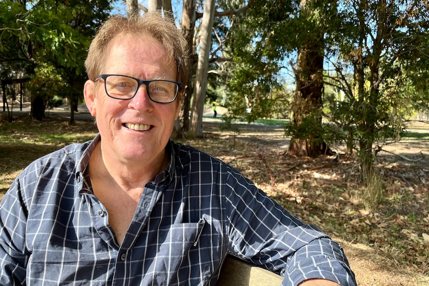 A man wearing a blue checkered shirt sits outdoors with trees and grass in the background.