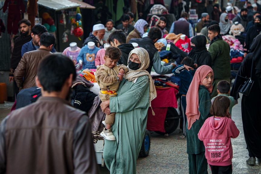 A woman walks through a market holding a small child, her hair and half of face is covered. She wears a long green gown.
