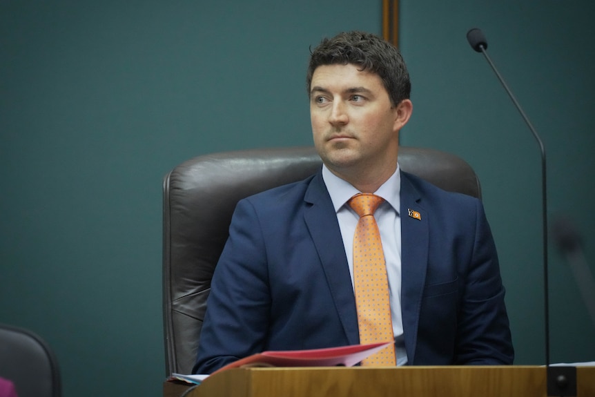 A white man, dark brown hair wearing a navy suit, orange tie sitting in a leather chair in front of teal wall.