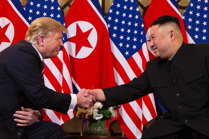 Donald Trump and Kim Jong Un shake hands while they sit in chairs in front of North Korean and US flags.
