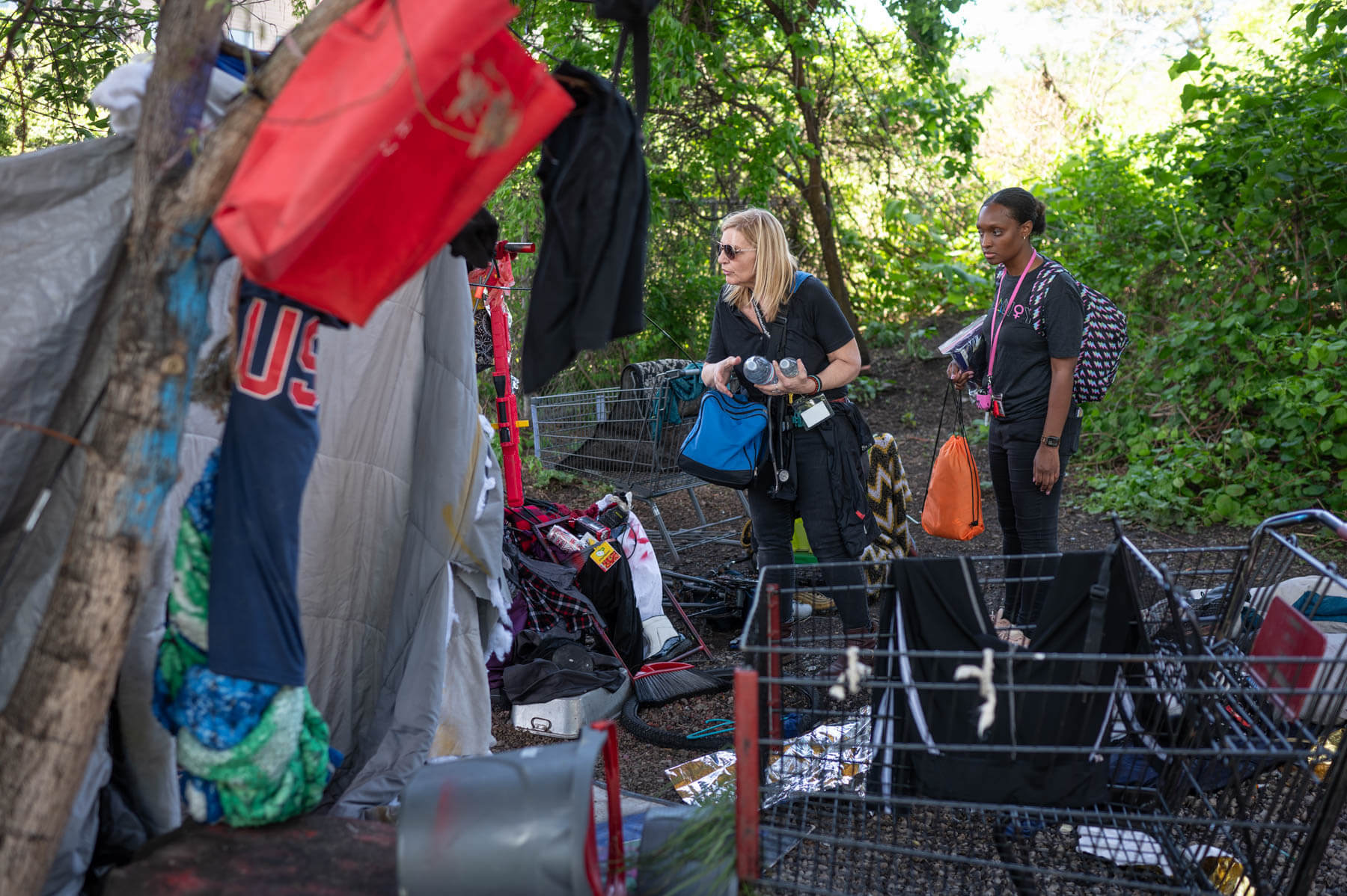 Two women observe items in an outdoor area with shopping carts, clothing, and various belongings, surrounded by greenery.