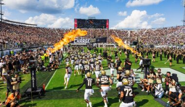 UCF football team wearing black uniforms, gold helmets and white pants charges onto field on party cloudy day