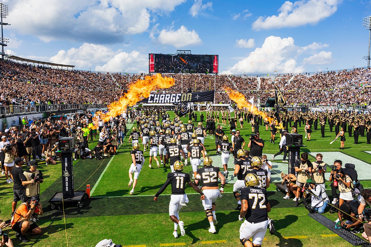 UCF football team wearing black uniforms, gold helmets and white pants charges onto field on party cloudy day