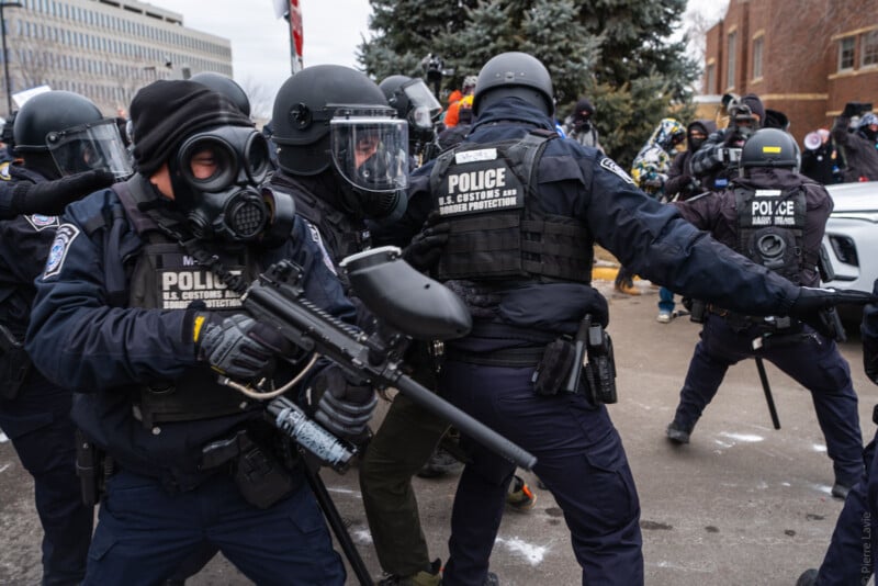 Police officers in riot gear and gas masks, labeled "U.S. Customs and Border Protection," push forward with batons and rifles during a tense outdoor confrontation. Other people are visible in the background near buildings and trees.