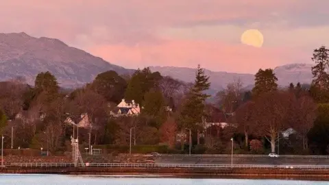 Sheila Quibell Buildings are partially obscured by trees which sit by a river. In the distance, a hill towers over the village. The sky has pink clouds and a large white moon 