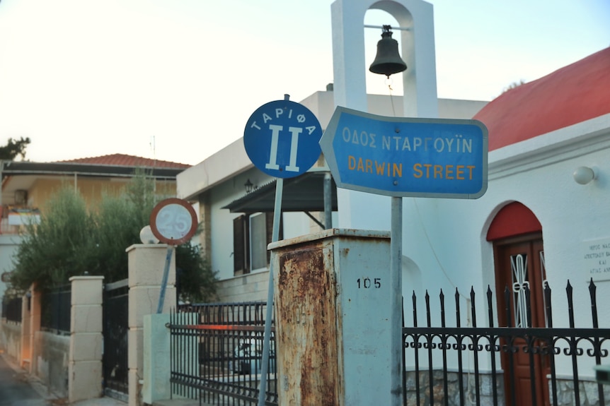 A blue street sign, partially in Greek with the other line reading 'Darwin Street' in yellow font, housing behind sign.