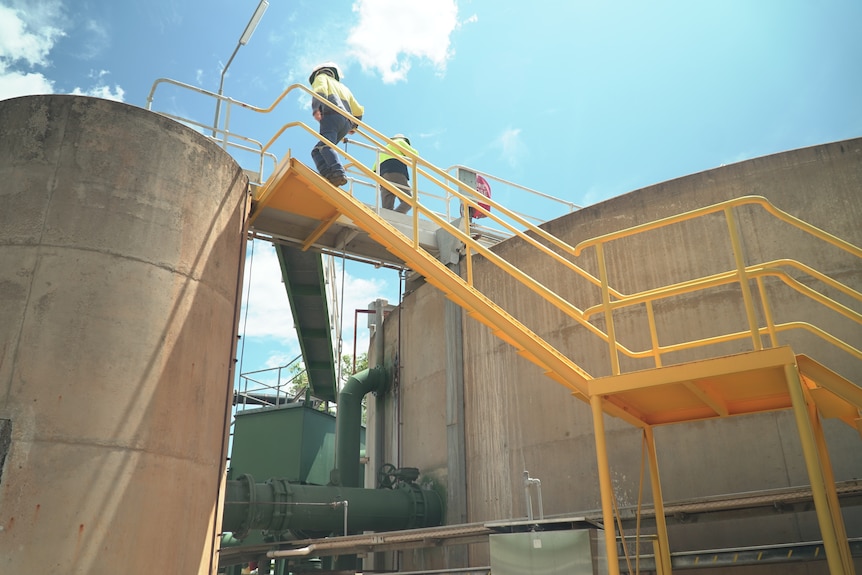 Two workers in high vis climbing stairs between two concrete tanks. 