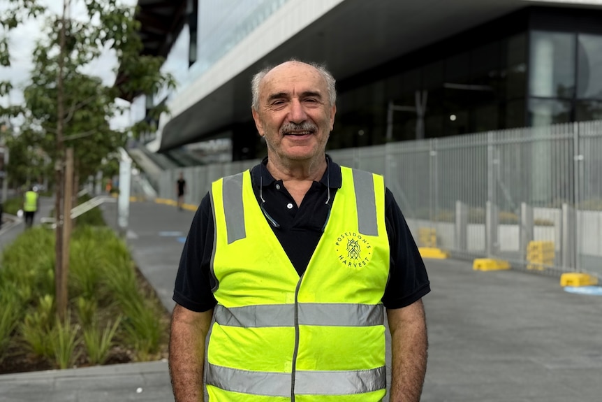 Kerry Strangas has short hair and a high-vis vest, and stands outside a large building.
