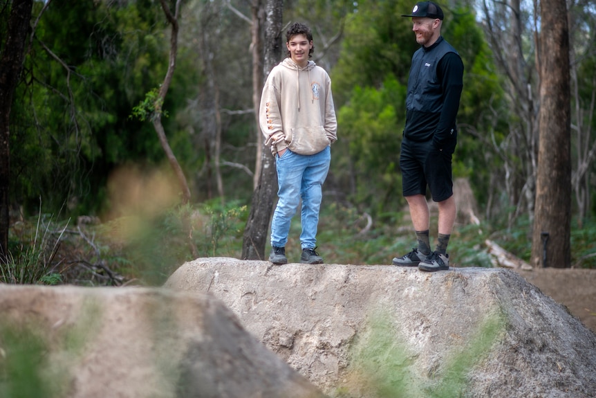 A man wearing a black cap smiles while chating with a young man in a beige hoody on top of steep dirt bike jumps in the bush