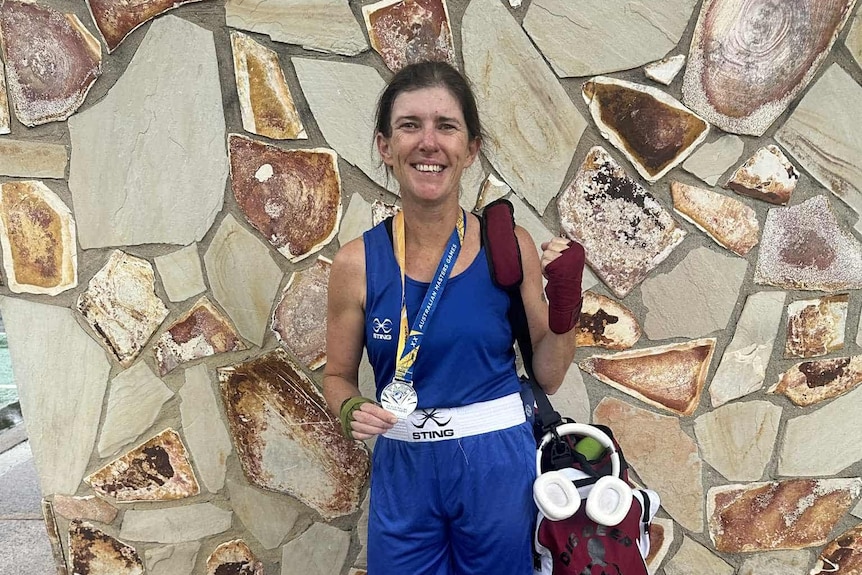 A woman in a blue boxing outfit, holding a medal and trophy and smiling.