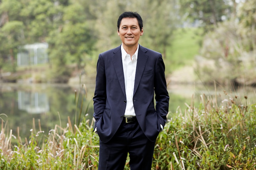 A man in a suit stands in front of a body of water in nature.
