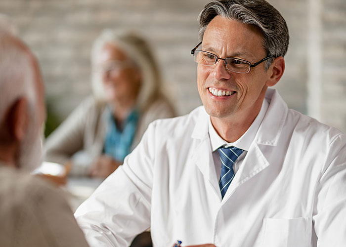 Male doctor wearing glasses and a white coat smiling and talking professionally to an elderly patient indoors