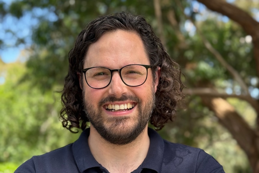 A smiling man with a beard in a blue shirt wearing glasses. 