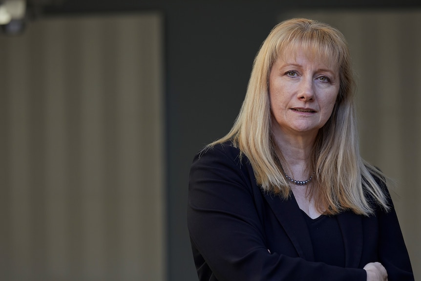 Bettina, a woman with blonde hair wearing a black jacket, looks seriously into the camera.