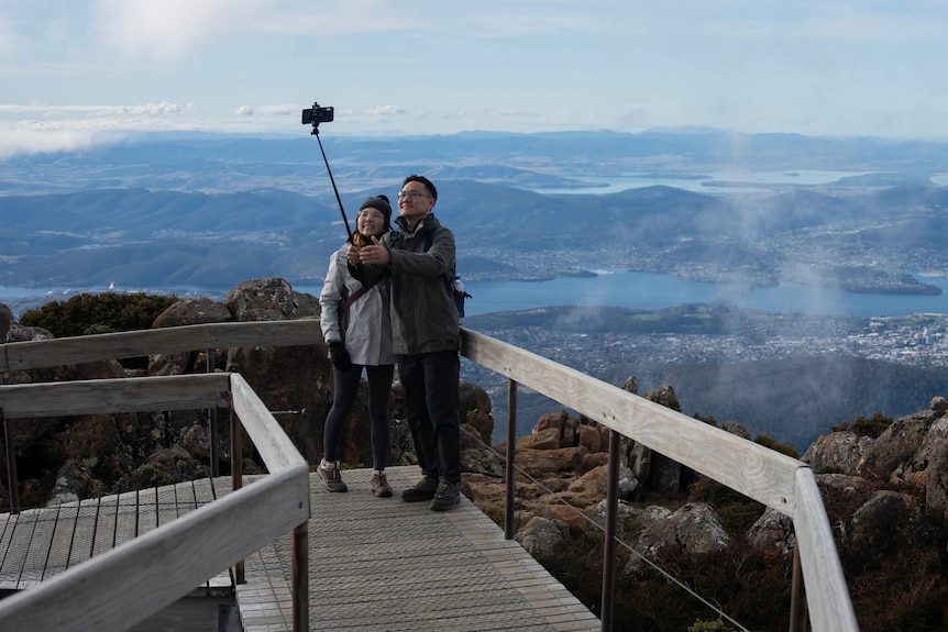 A couple stand on a boardwalk taking a pic with a selfie stick with Hobart landscape behind