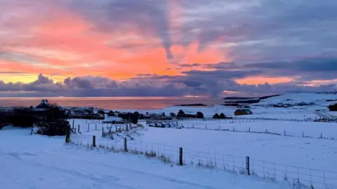 BBC Weather Watcher/Wendy a blanket of snow over fields with water in the distance and a pink/orange sunrise 