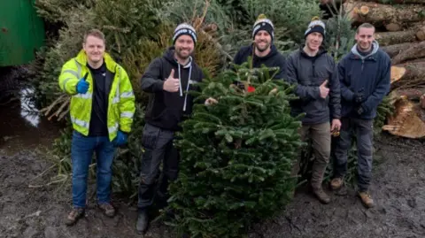 Ringwood Round Table Five men wearing outdoor clothing, some giving thumbs-up signs, stand by piles of Christmas trees and logs.