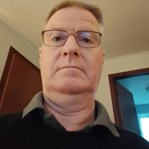 Craig Mitchell A man with short, light brown, greying hair and glasses. He has a green/grey T-shirt on with the collar sticking out from under a dark jumper.