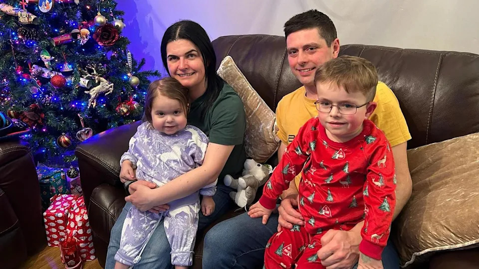 A family of four sit on on a brown leather sofa by a Christmas tree. The mother with medium length dark hair holds her daughter who is wearing purple unicorn pajamas on her knee. The father, with short brown hair, wearing a yellow t shirt, hold his son, who is wearing red Christmas tree pajamas.