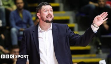 Marc Steutel, wearing a suit with an open-necked white shirt, makes a gesture with his left hand towards his basketball players