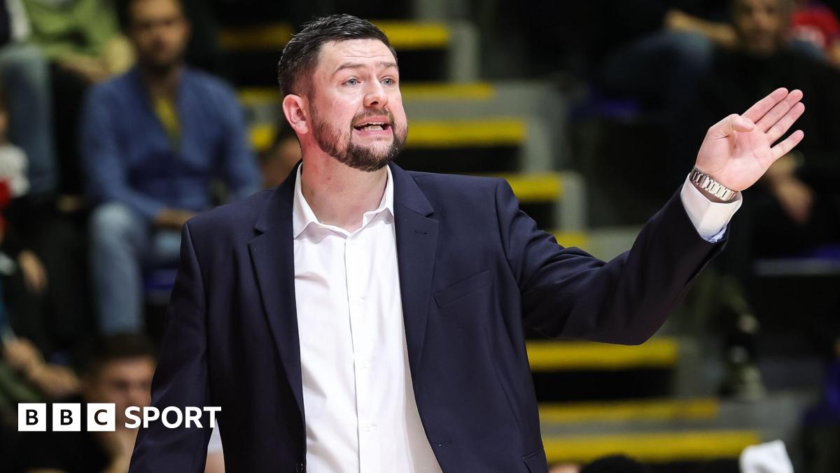 Marc Steutel, wearing a suit with an open-necked white shirt, makes a gesture with his left hand towards his basketball players