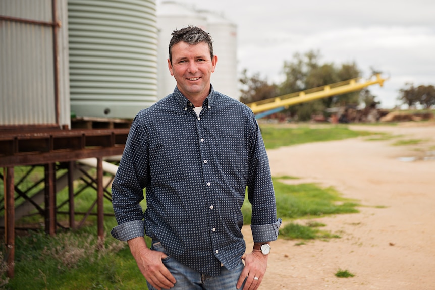 Man with white skin and blue shirt stands on rural property in front of water tank