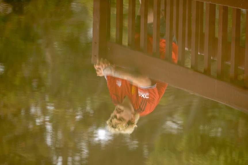 The reflection of a man in water, standing at the railing of a bridge