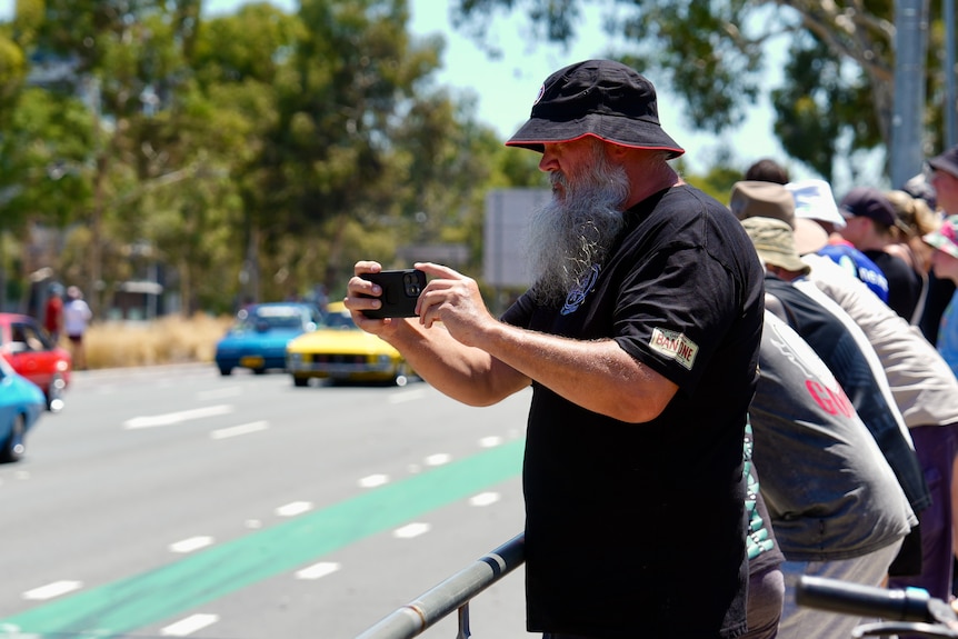 A man takes a photo on his phone of a car parade