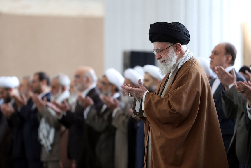 A photo of a man wearing a brown tunic, black turban. He's holding his hands up in prayer. Nine men are lined behind him.